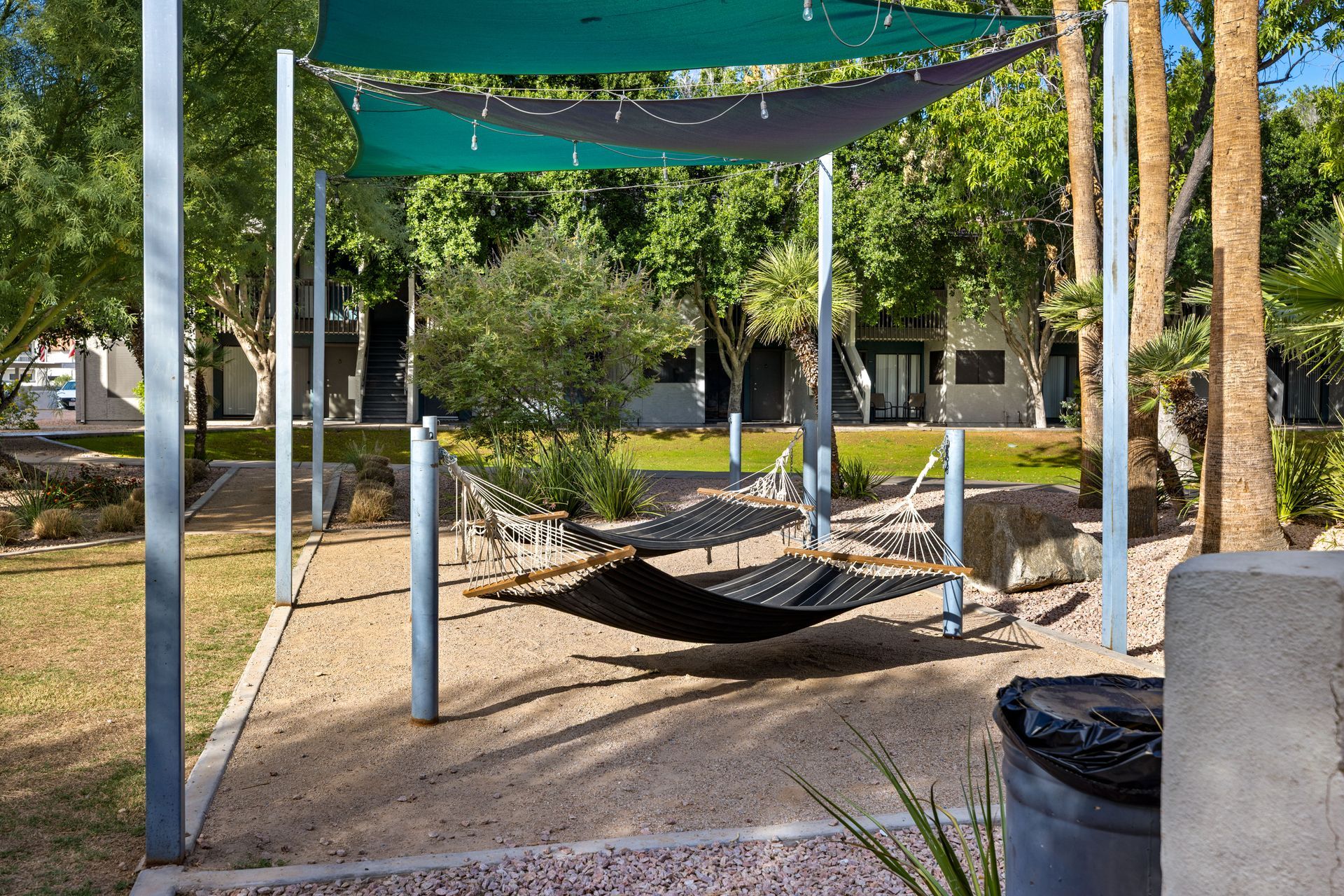 Hammock under a shade structure in a park-like setting. Trees and buildings are in the background.