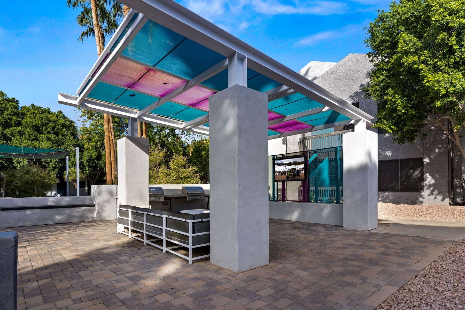 Outdoor kitchen with a colorful, shaded canopy over a grill area and seating on a brick patio.