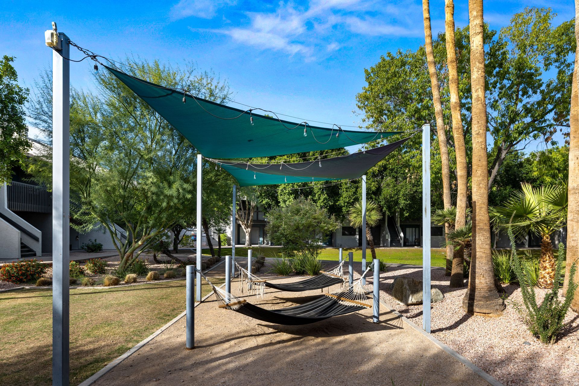 Shade sails over hammocks in a park setting with palm trees and a blue sky.