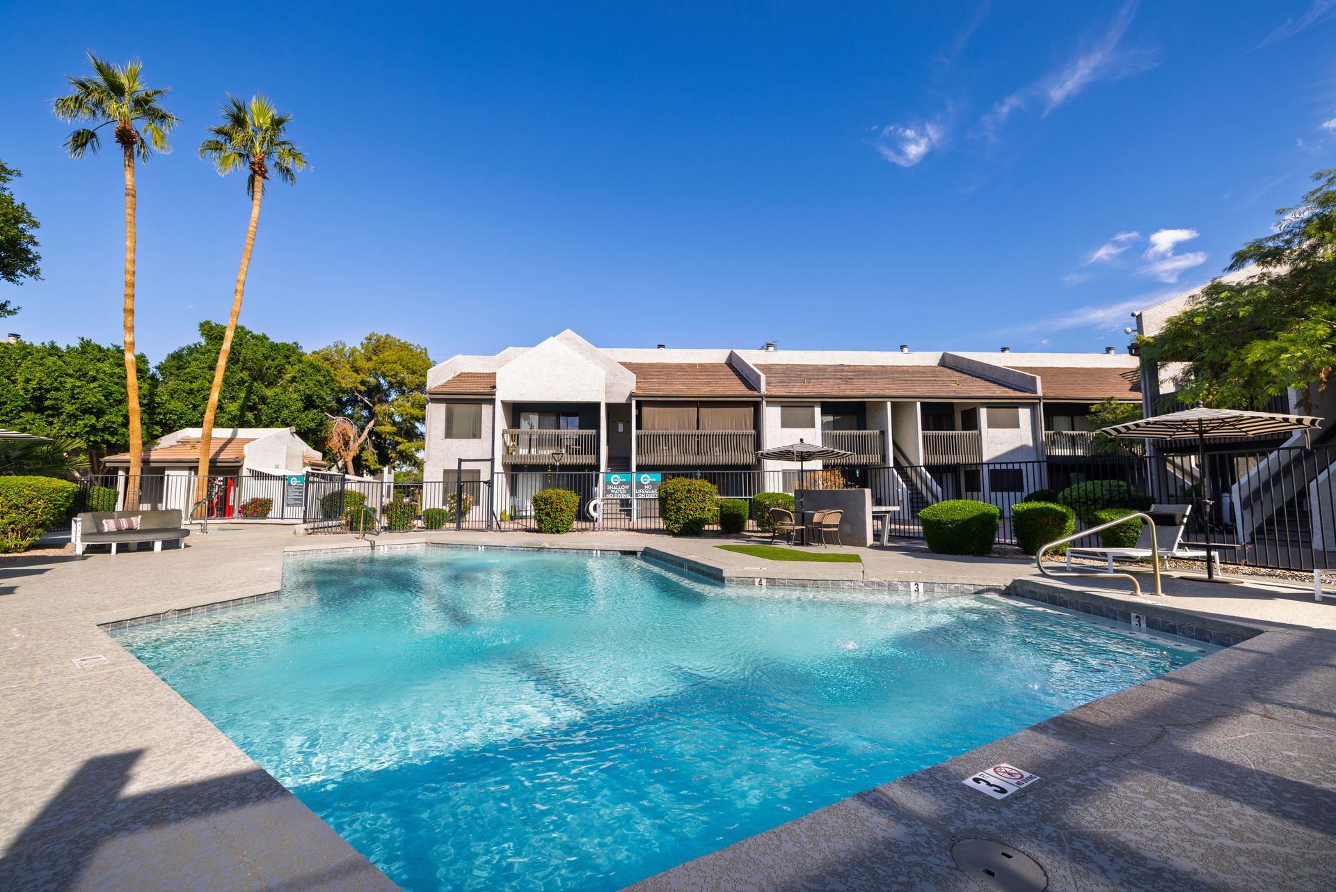 Pool in front of a two-story apartment building under a blue sky, palm trees.