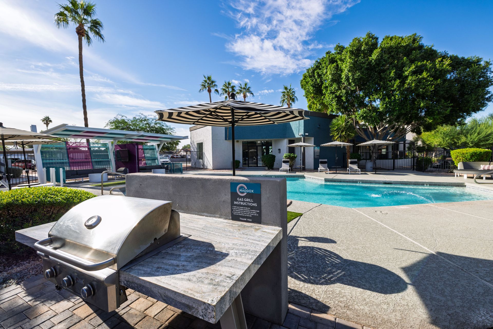 Outdoor grilling area near a swimming pool with palm trees under a blue sky.