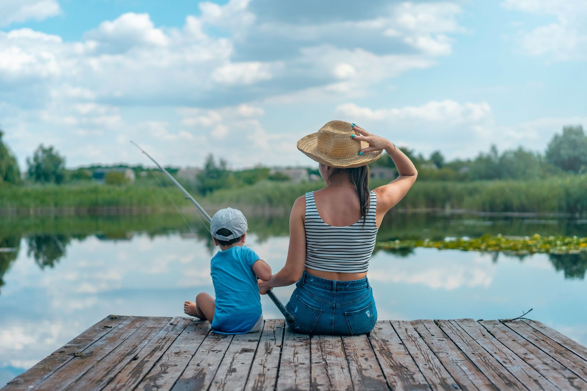 A person and a child sit on a wooden pier, holding a fishing rod over a calm lake under a cloudy blue sky.