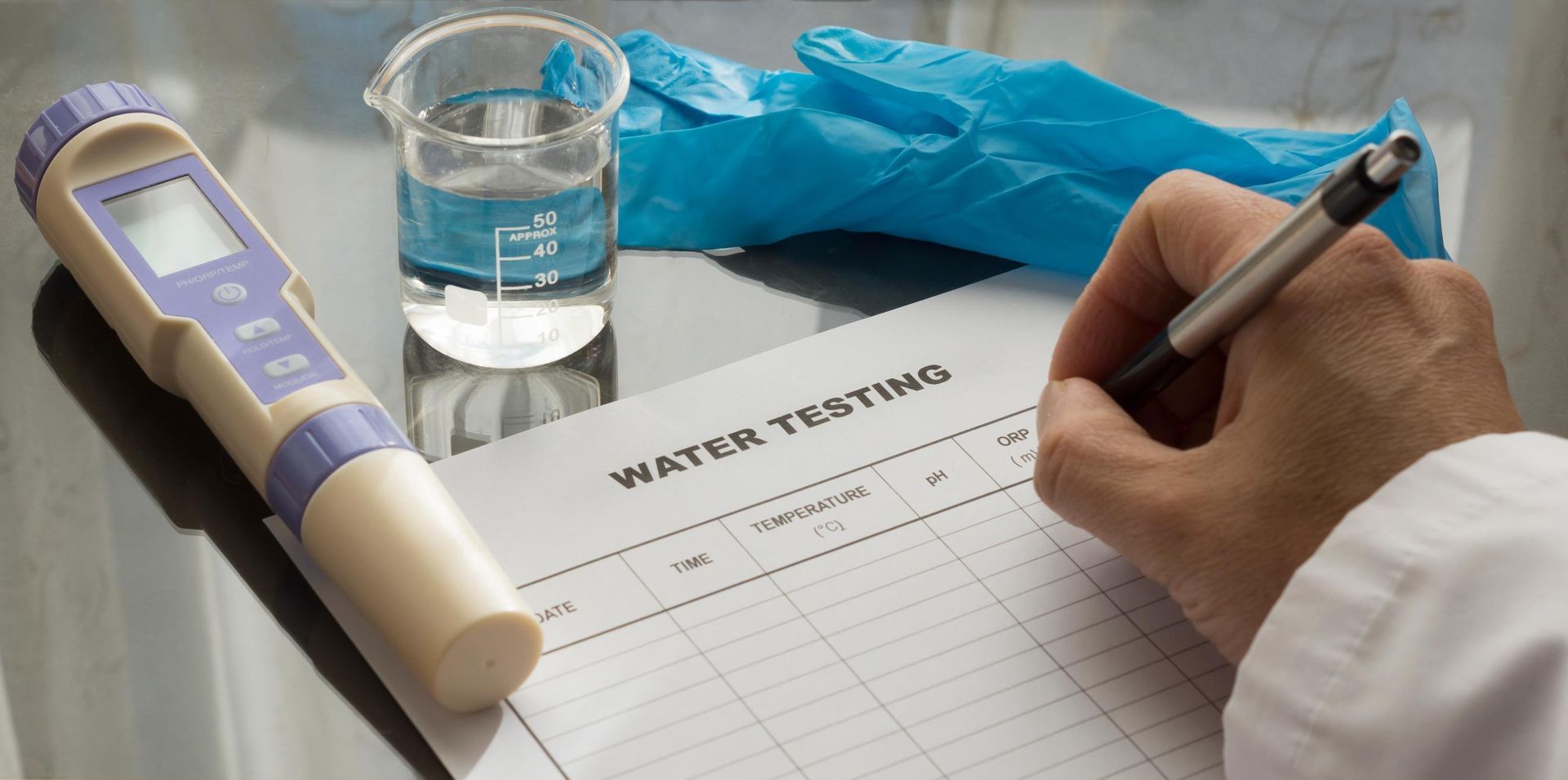 A person tests water quality, using a meter, beaker, and gloves, and records results on a