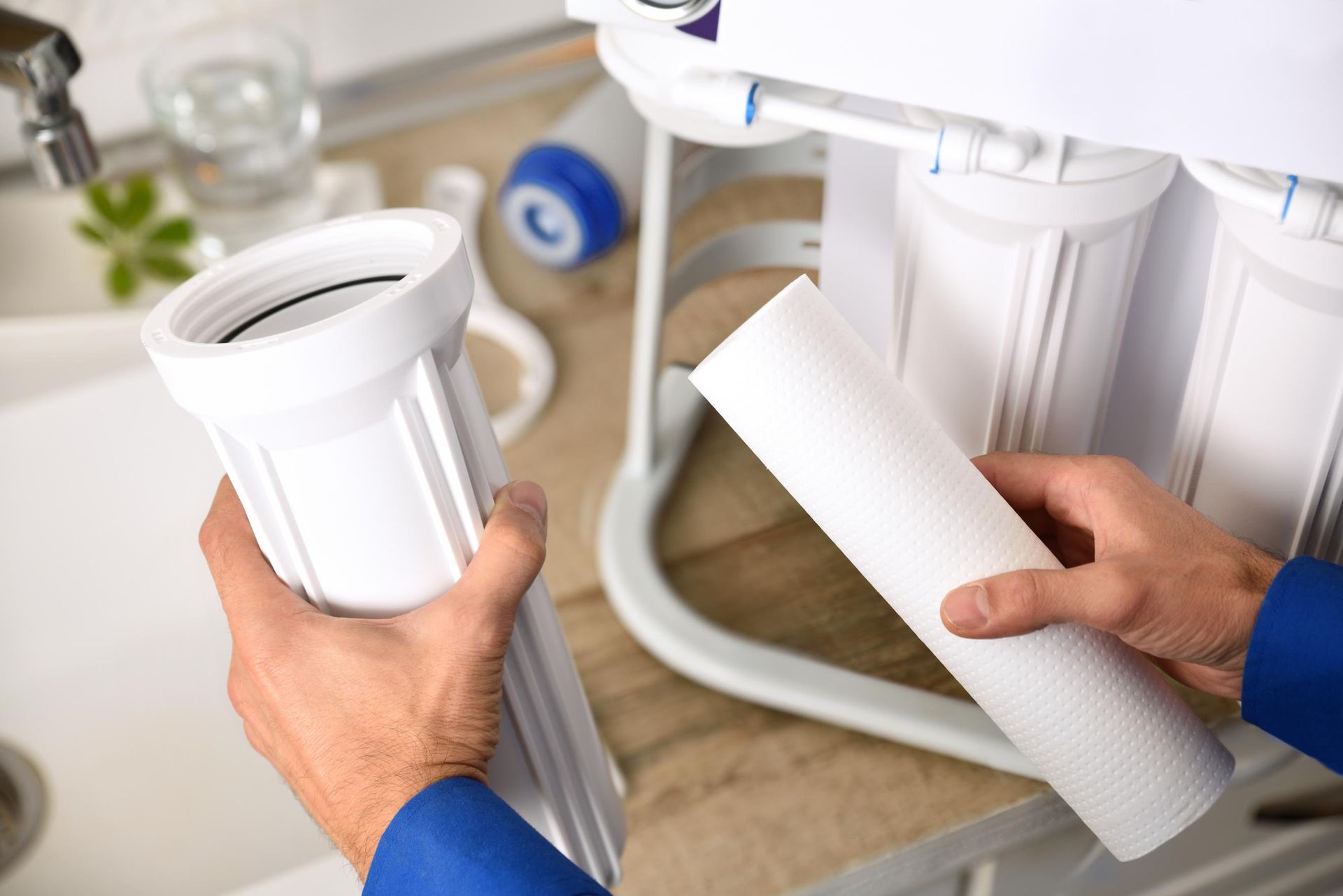Hands holding a white water filter and housing, replacing a filter in a reverse osmosis water filtration system in a kitchen.
