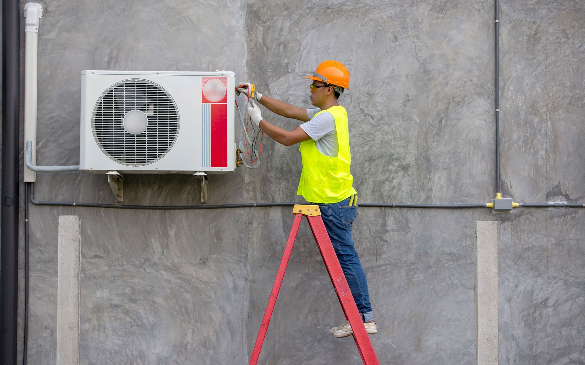 A man is standing on a ladder fixing an air conditioner on the side of a building.