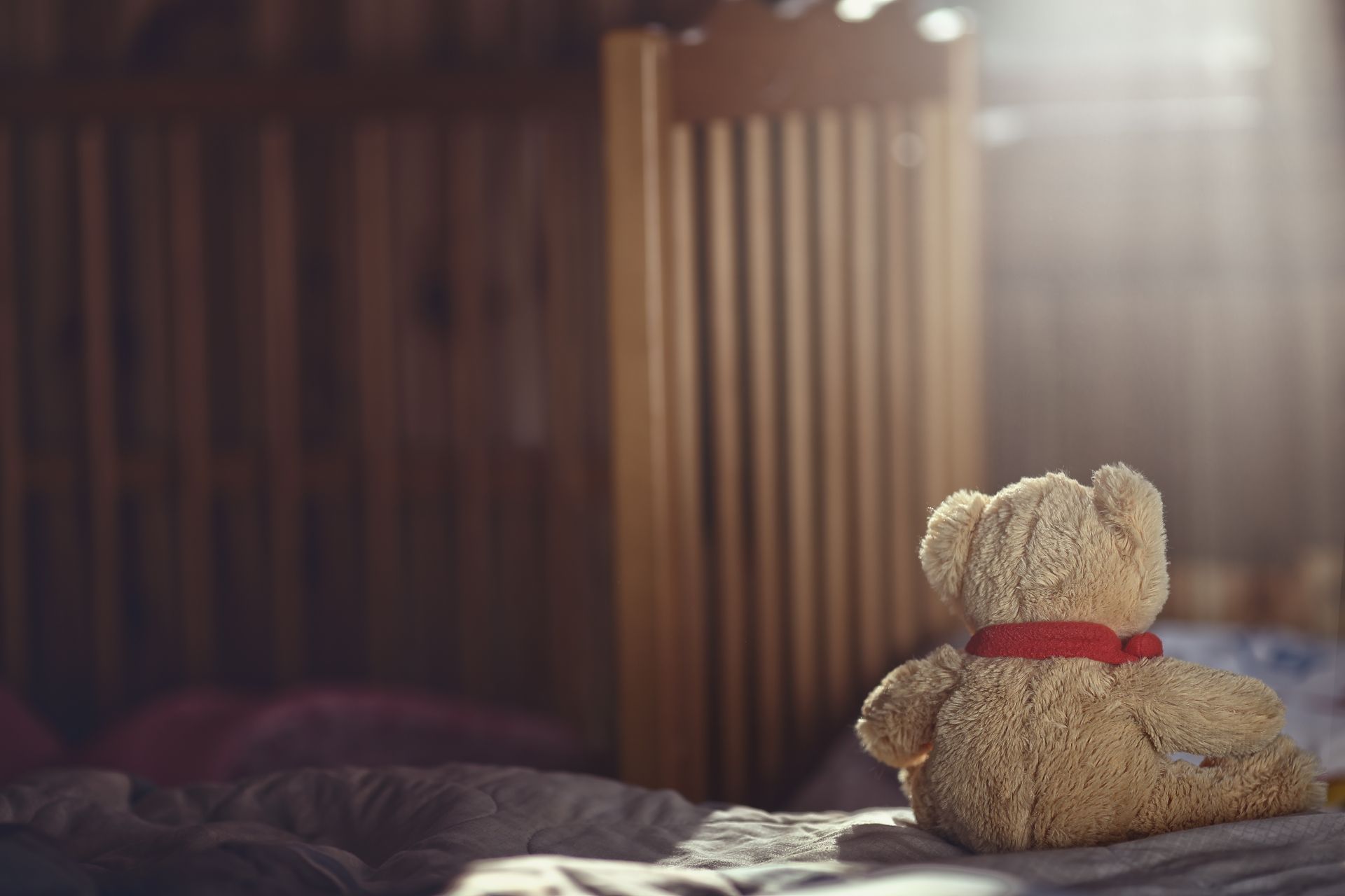 A small, tan teddy bear with a red collar sits on a rumpled bed, facing a wooden crib in a sunlit room.