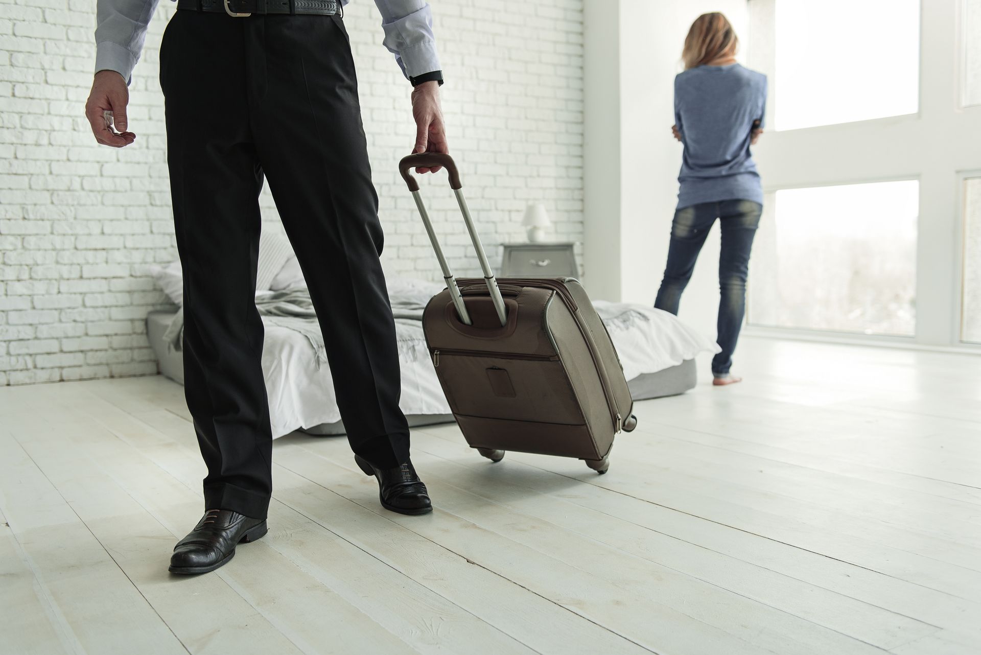 A person walks away with a suitcase while another stands in the background of a sparse room.