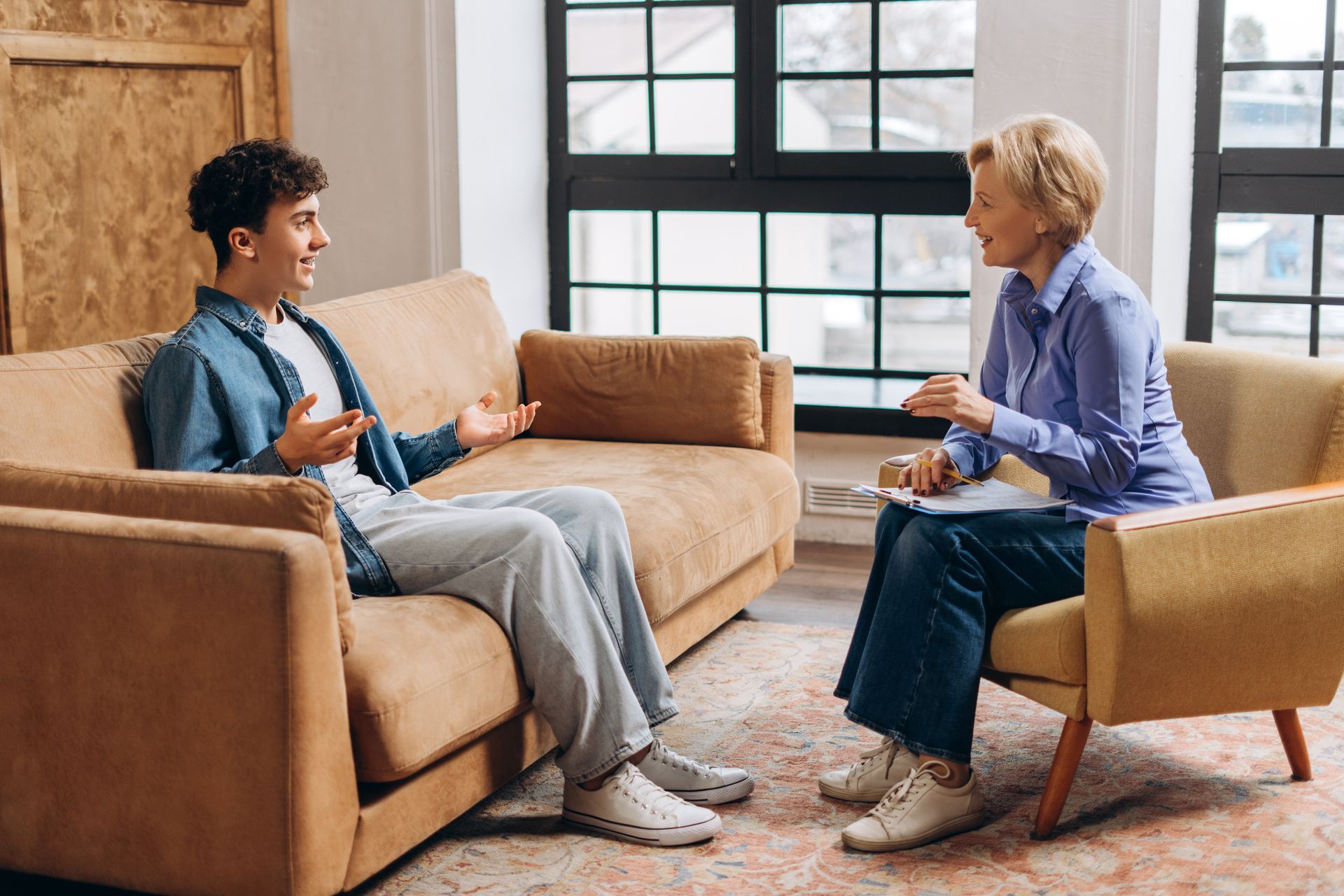 Man on couch speaking to person in armchair holding notepad; indoors, near window.
