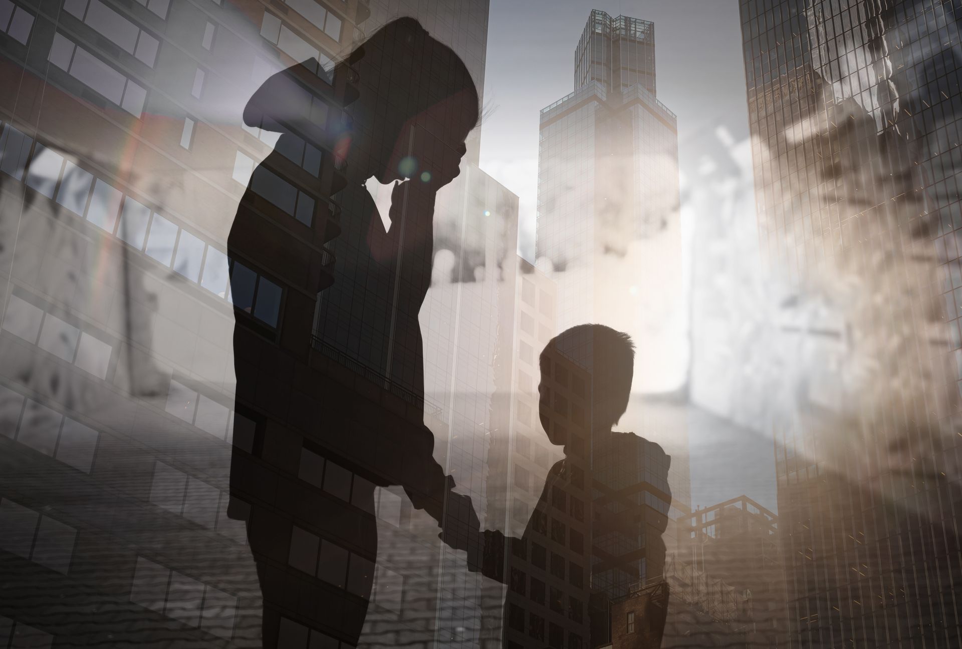 A silhouette of an adult holding the hand of a child against a double-exposed background of a city street and buildings.
