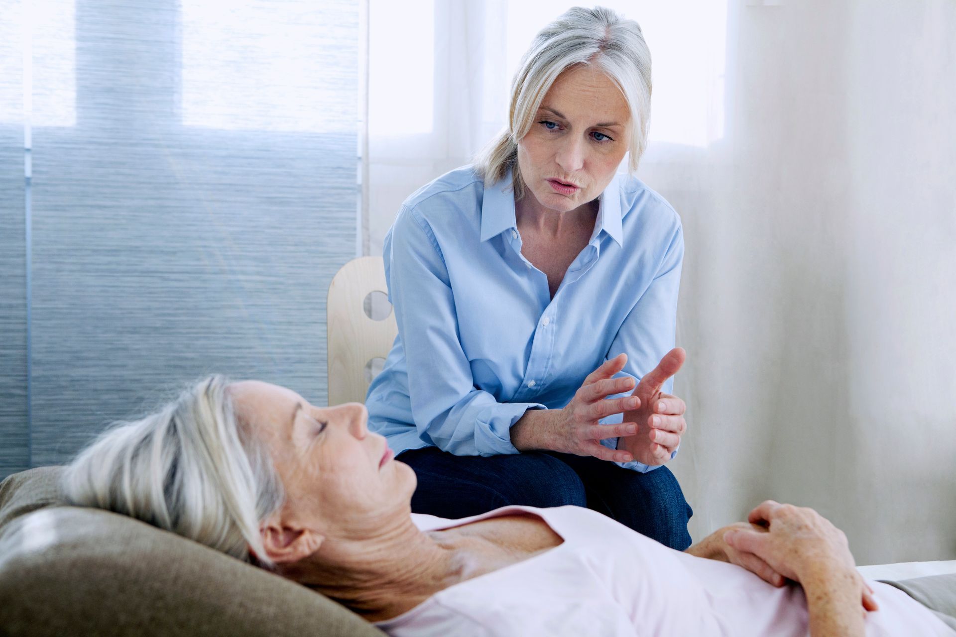 Woman in blue shirt comforting an older person lying in bed in a bright room
