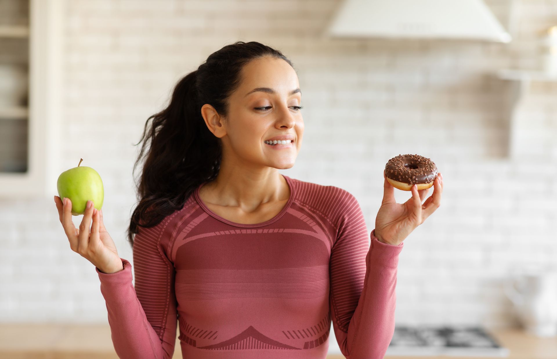 A person in athletic wear holding a green apple in one hand and a chocolate donut in the other, smiling in a kitchen.