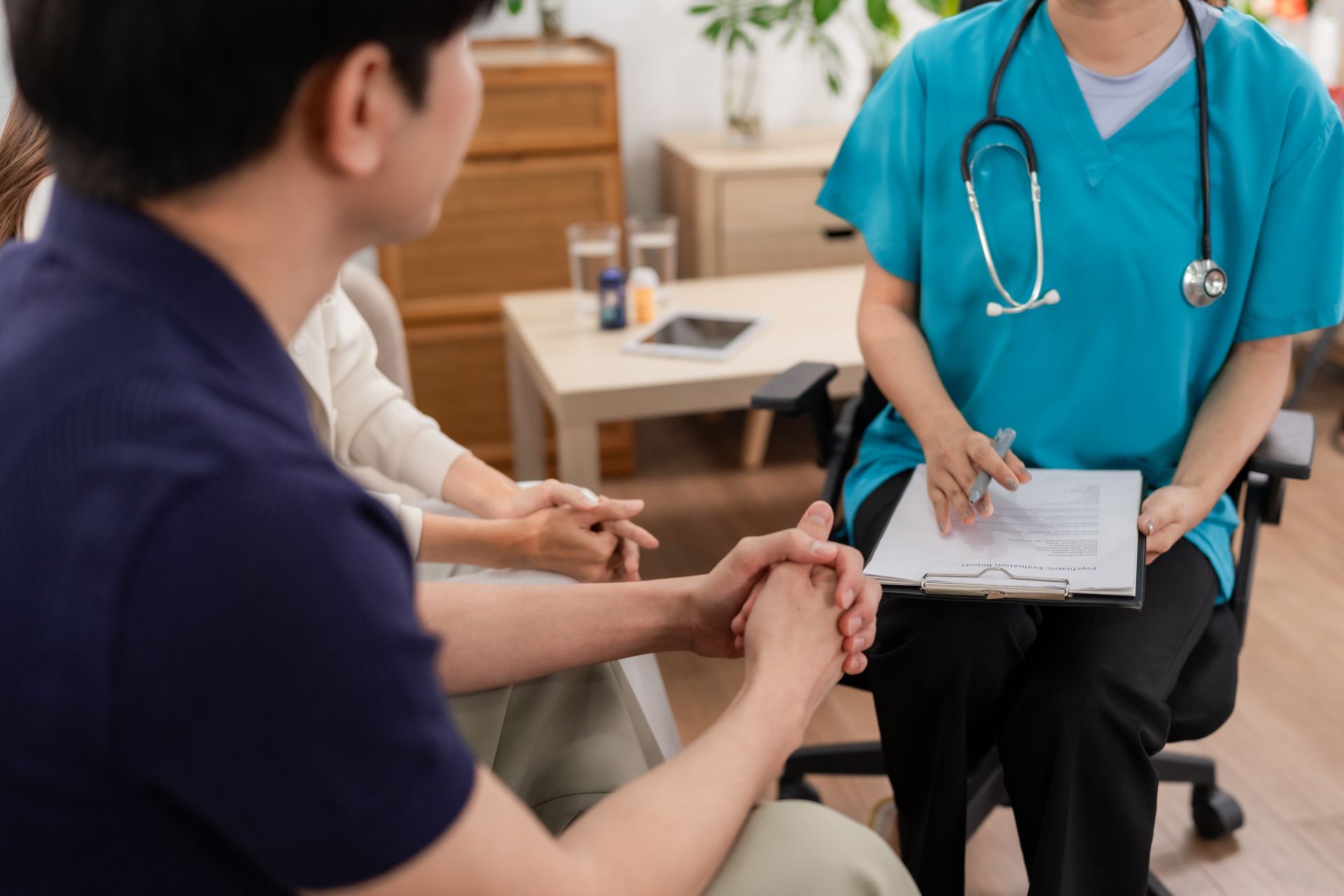 Couple consults with a medical professional, indoors; patient holds hands, doctor notes on clipboard.