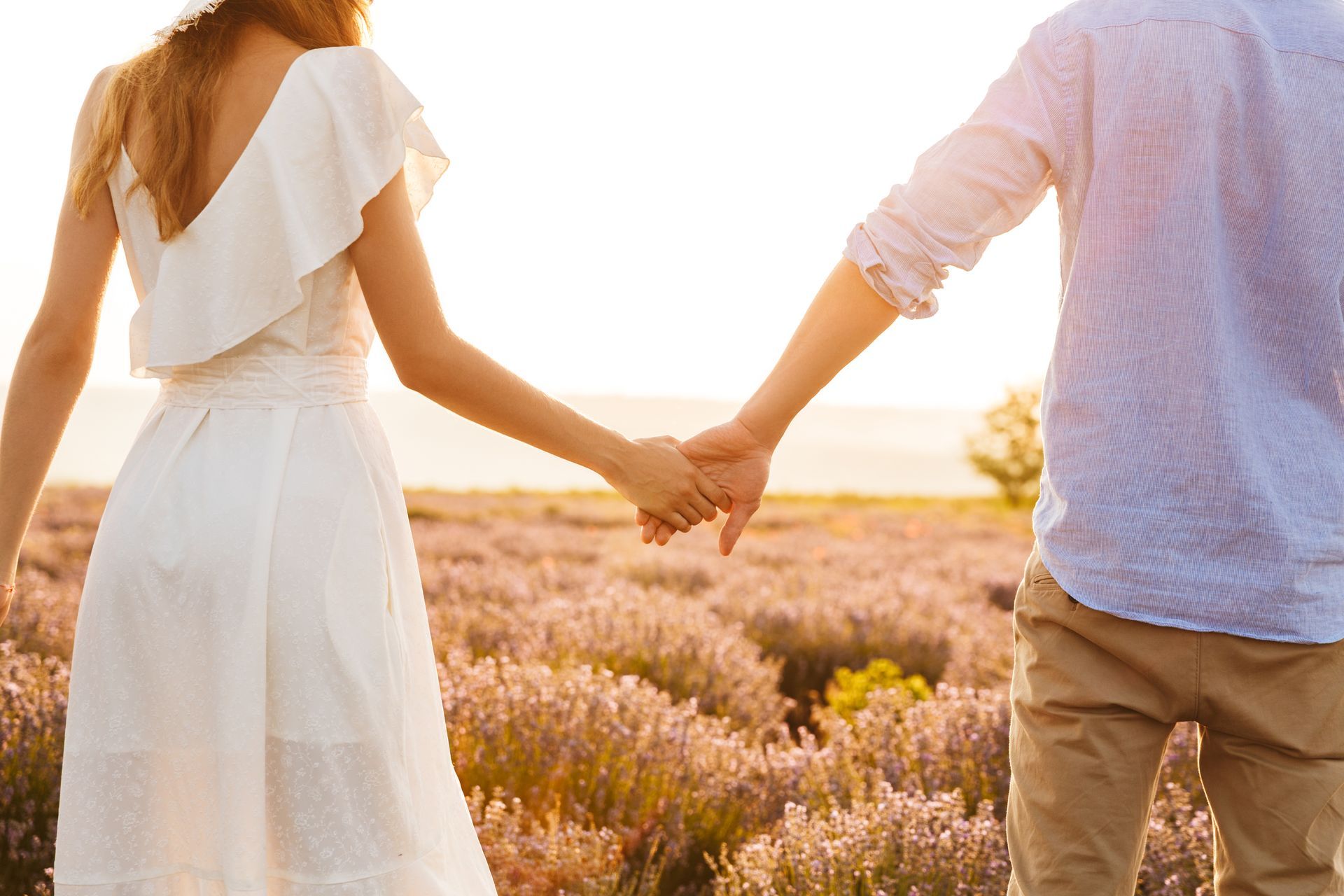 A person in a white dress and a person in a light blue shirt hold hands while walking through a blooming lavender field.