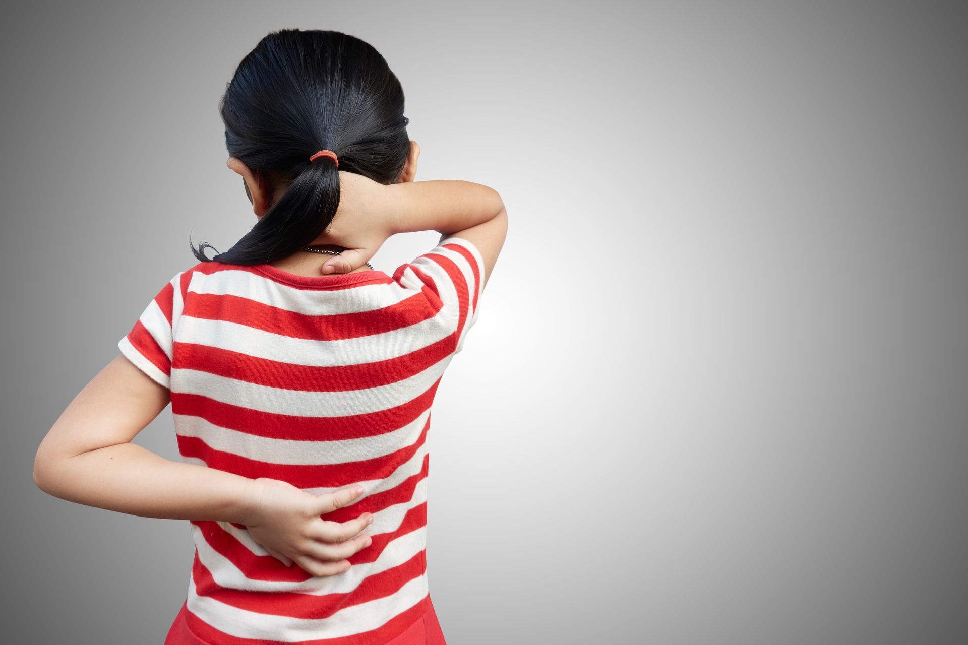 Person in red-and-white striped shirt touching neck and lower back, viewed from behind on gray background