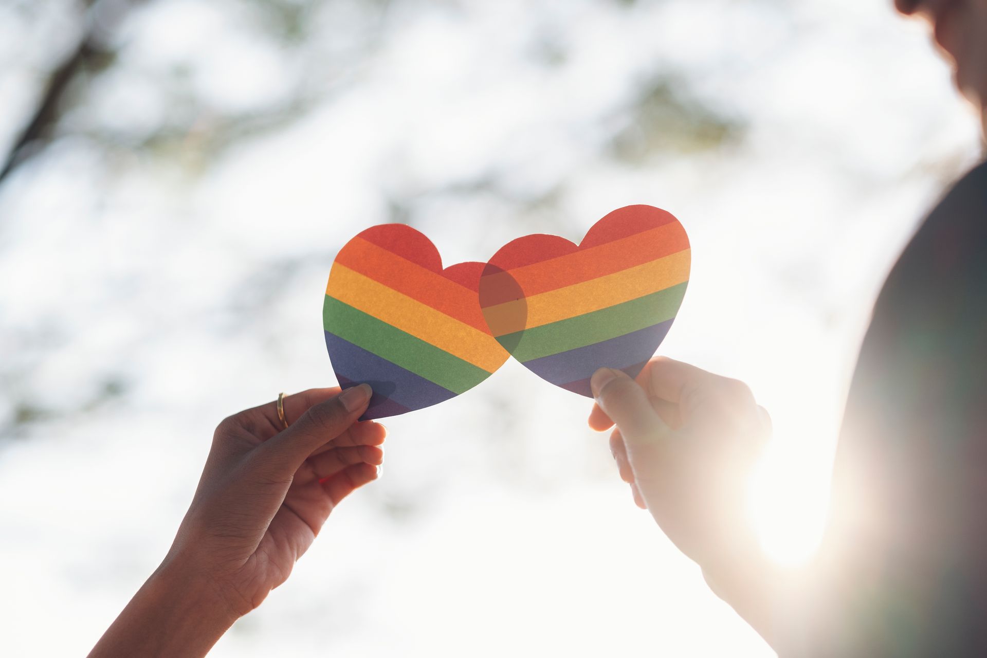 Two hands held up to the sky, each holding a rainbow-striped heart cutout that overlap against a soft, sunny background.
