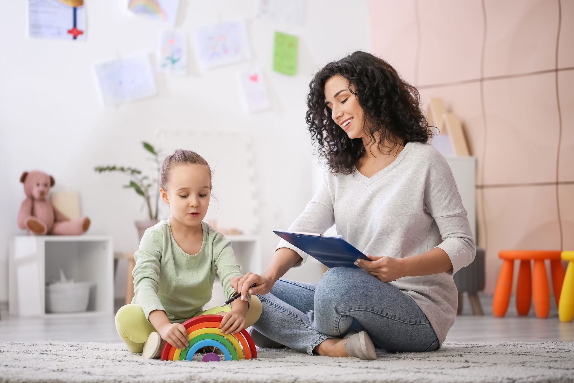 Woman and child playing with rainbow toy on floor. Woman holds clipboard, smiling.