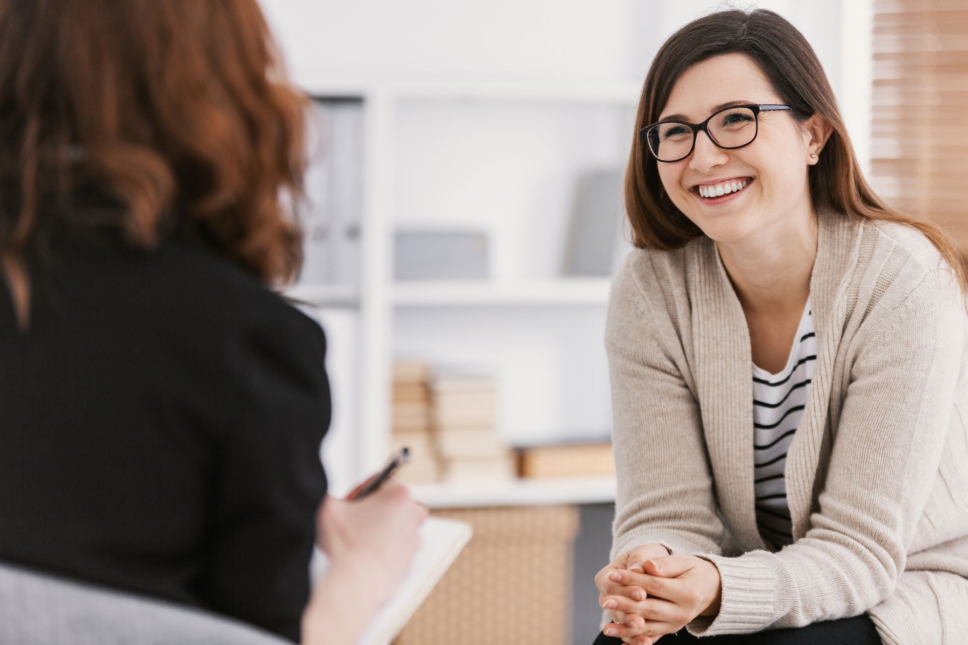 Woman smiling in therapy session, facing a therapist who is taking notes. Bright, airy room.