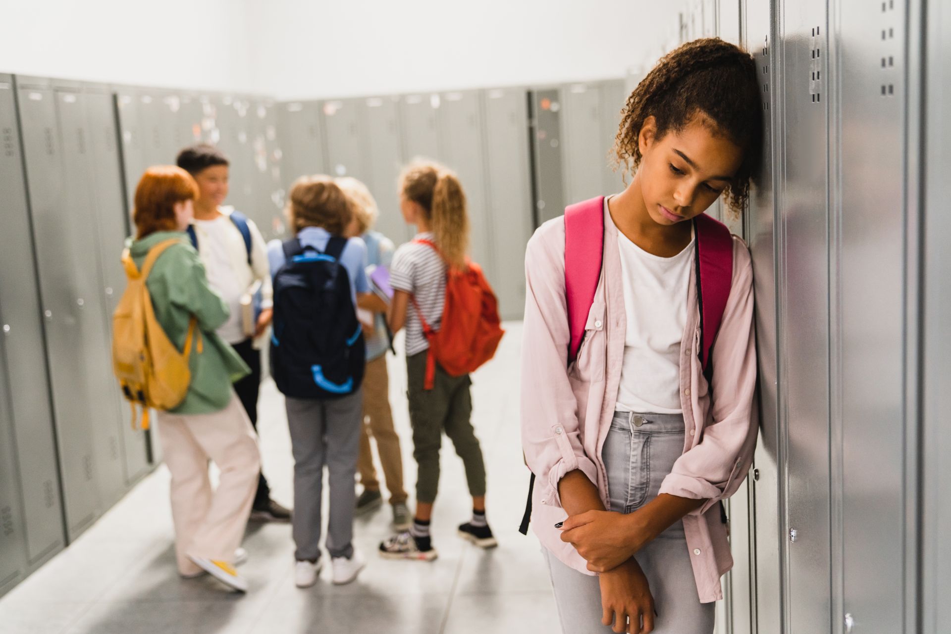 A student leans against school lockers with their head down, looking sad, while a group of students talks in the background.