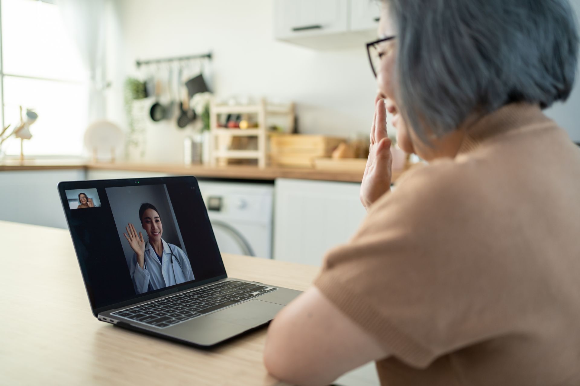 Woman in a telehealth session waving at a doctor on a laptop in a kitchen setting.