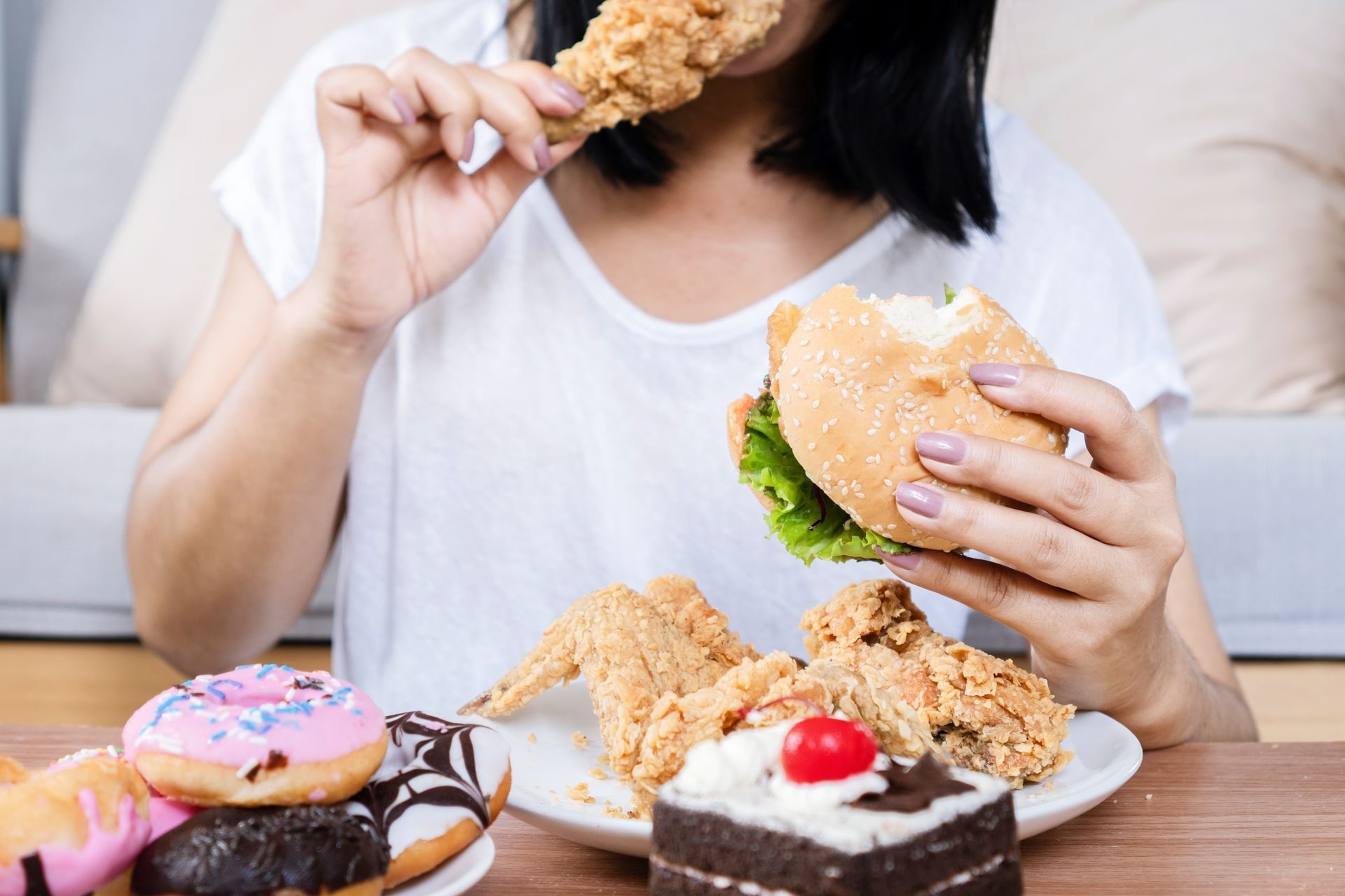 Person eating fried chicken and a burger at a table with donuts and cake。
