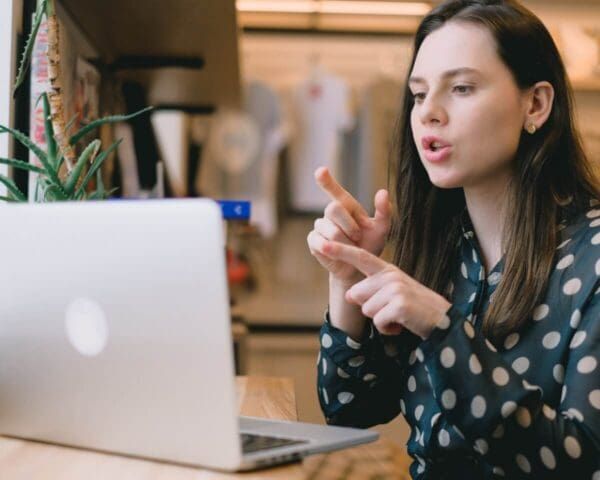 A person in a polka-dot shirt sits at a desk using sign language while video calling on a laptop.