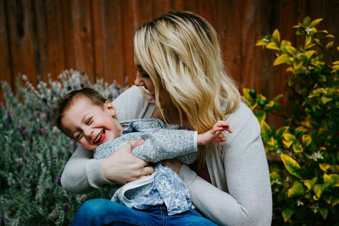 Woman in gray sweater holding a smiling child outdoors in a garden.