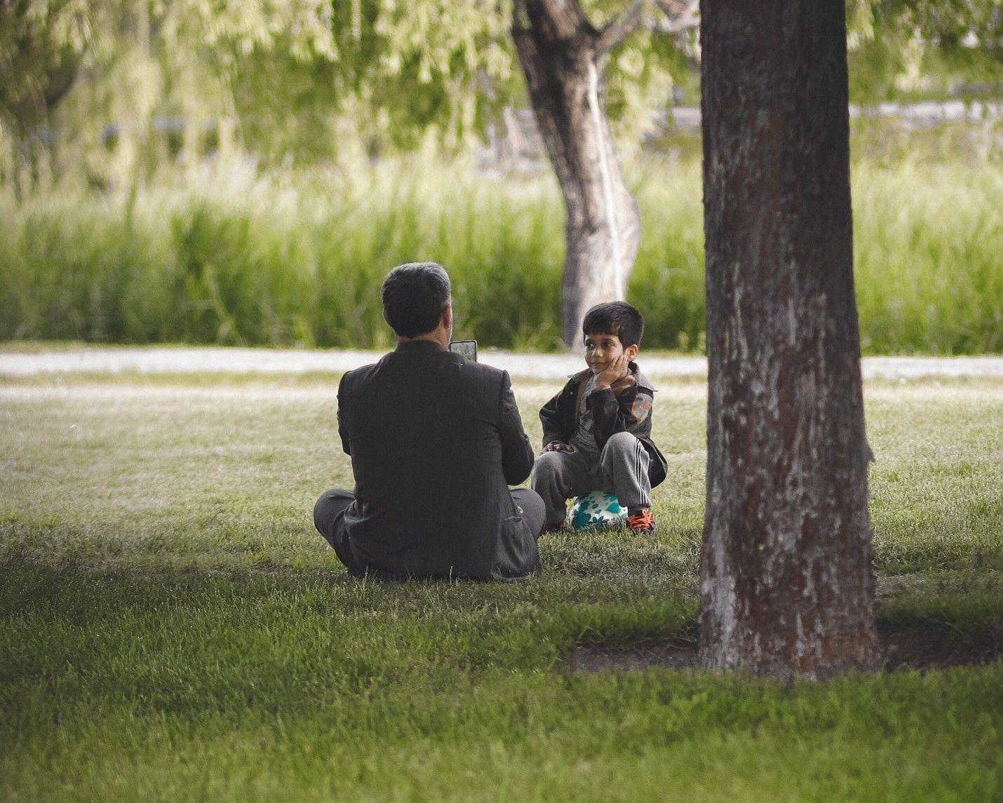 A person in a dark suit sits on a grassy lawn facing a young child sitting nearby under the shade of a tree.