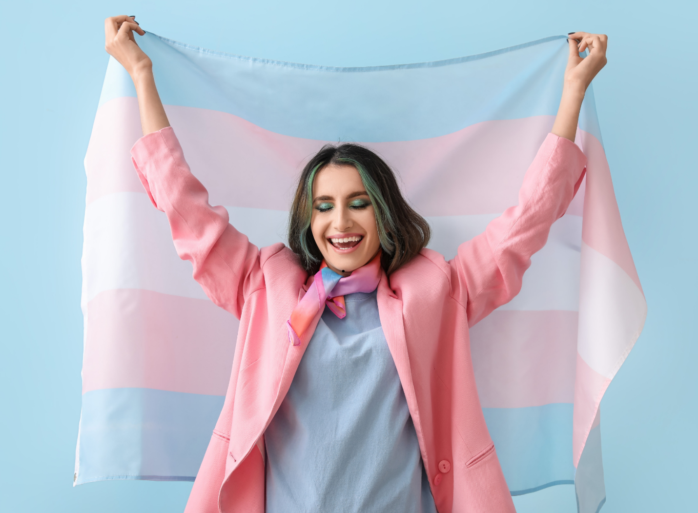 Smiling person in pink jacket holding a pastel blue and pink flag with arms raised
