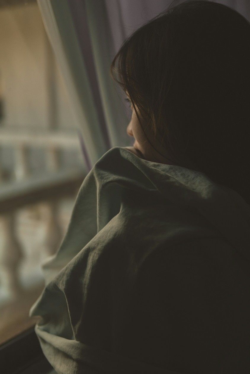 Person wrapped in a green blanket gazes out a window in soft light