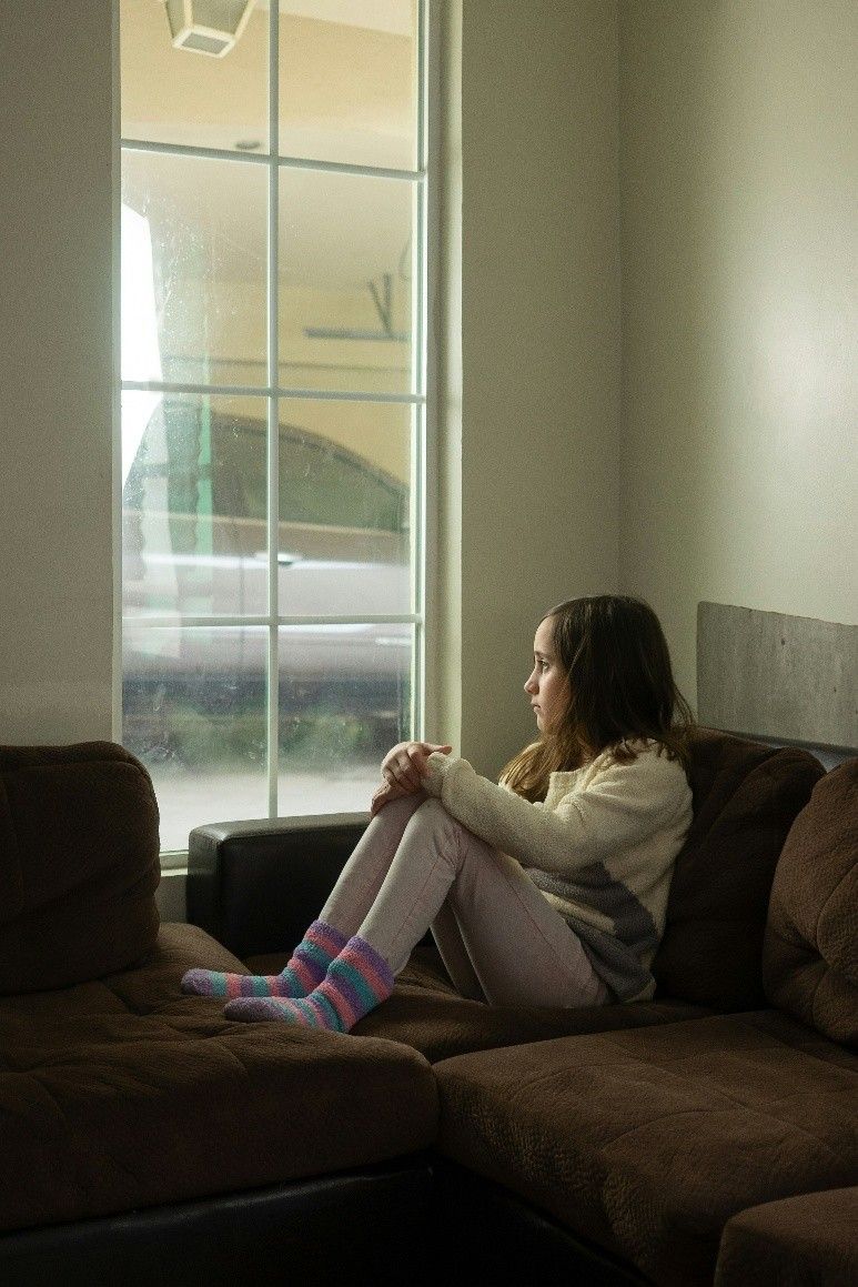 Person sitting on a brown couch by a window, looking outside with knees hugged to chest.