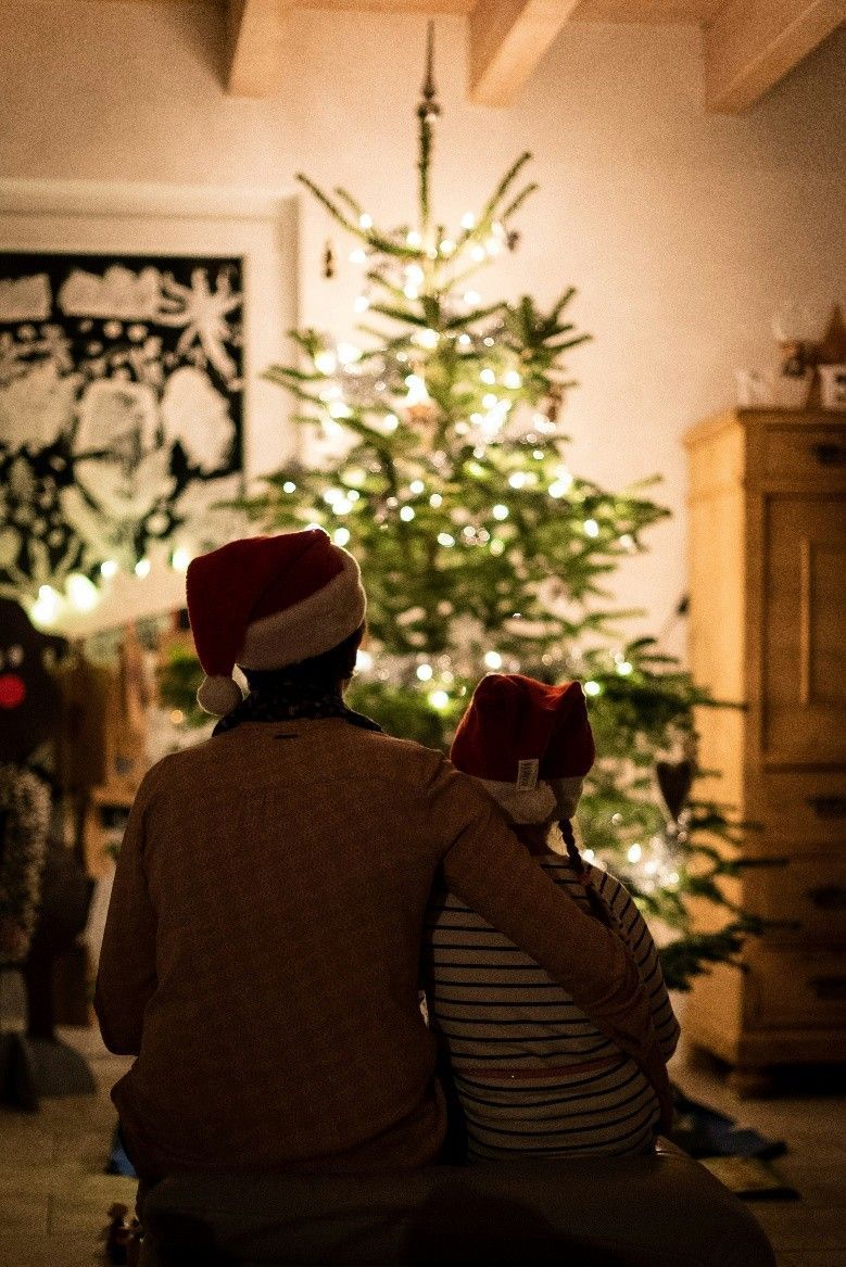 Two people in Santa hats sit by a lit Christmas tree in a cozy living room