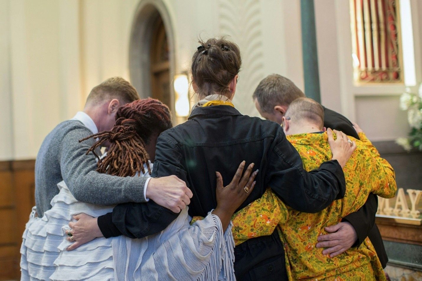 Four people stand arm-in-arm in a quiet indoor circle, wearing patterned clothing and jackets.
