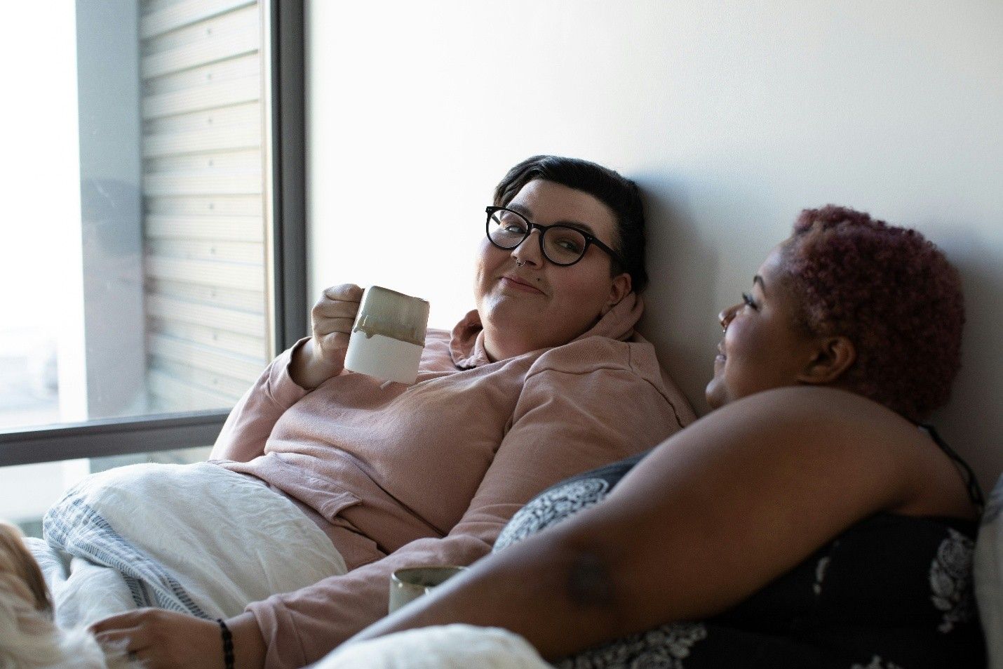 Two people sitting on a couch, one holding a mug and smiling in a bright living room.