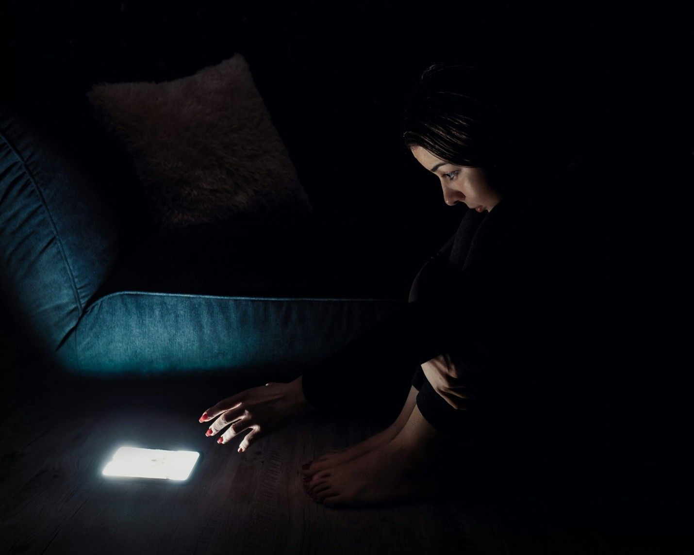 Person sitting in a dark room, reaching toward a glowing phone on the floor near a couch.
