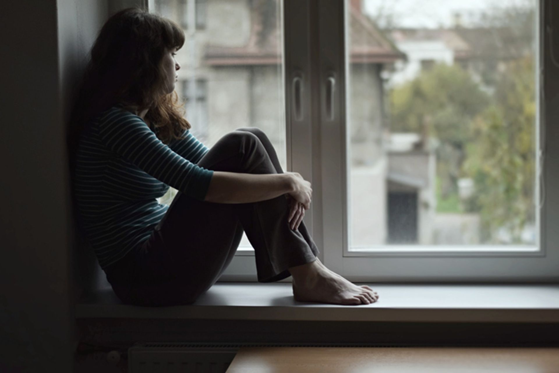 Person sitting curled up on a windowsill, looking out at a suburban street.