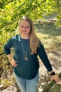 A person with long blonde hair smiles while standing outdoors in a teal long-sleeve shirt, jeans, and a pendant necklace.