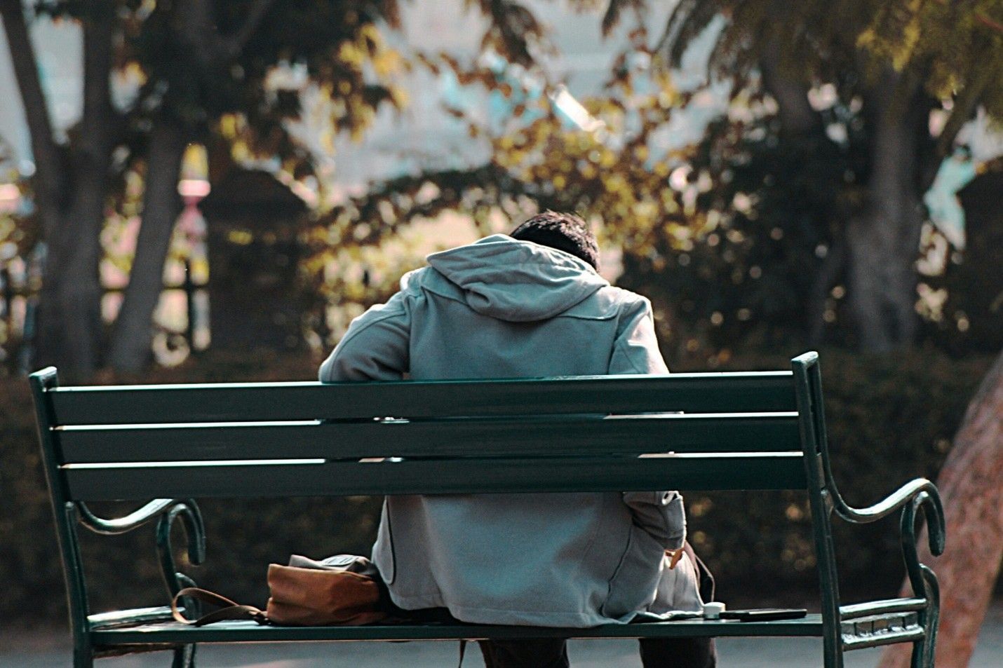 Person in a green hoodie sits on a park bench, seen from behind, with a bag beside them.