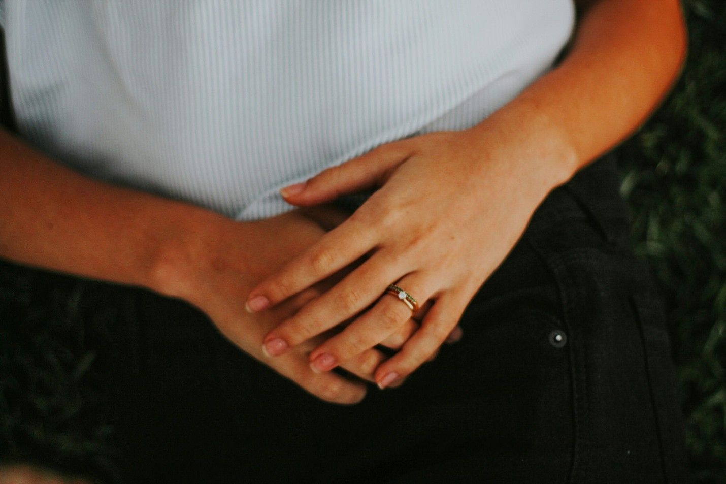 A person’s hands rest on their lap, wearing a simple gold band on their ring finger, set against dark fabric and greenery.