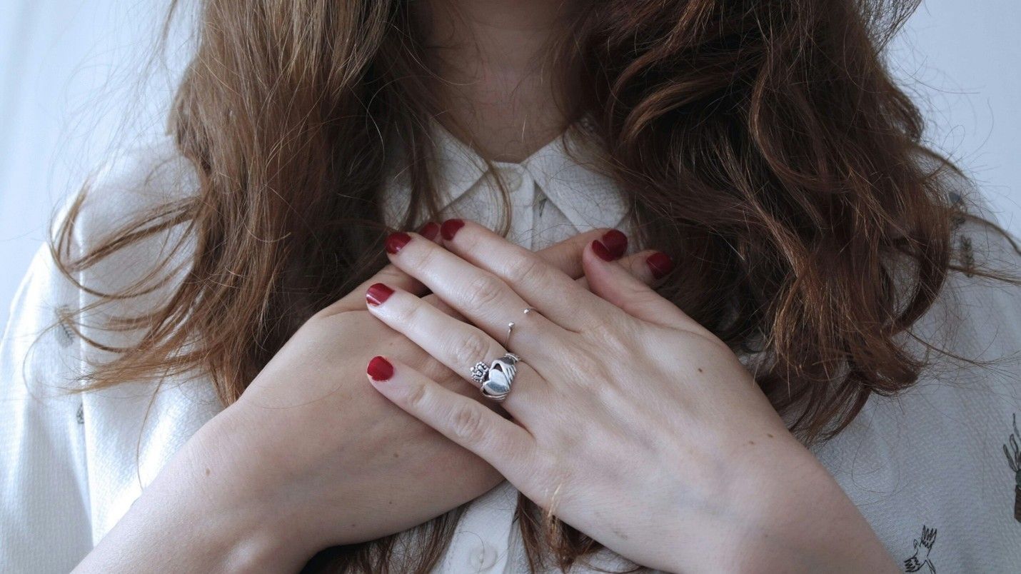 Hands with red nail polish and a ring resting on a white blouse with a braided tie.