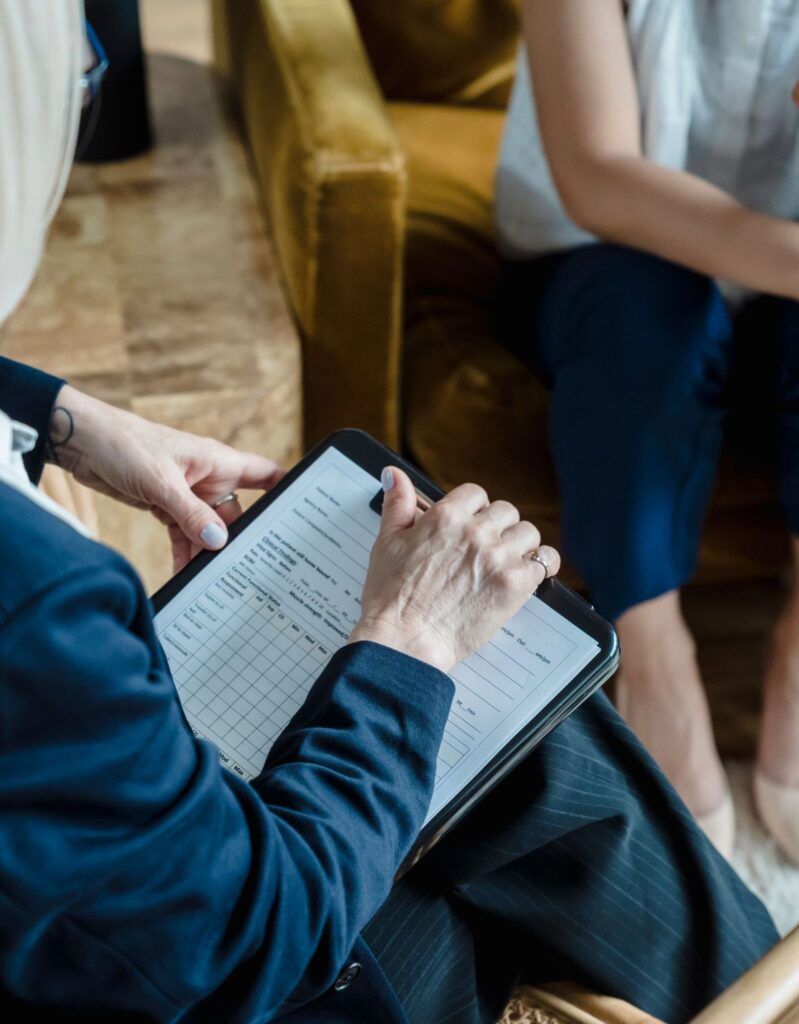 A person in a blue blazer holds a clipboard with a grid, sitting opposite another person in a professional setting.