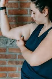 A person wearing a dark tank top pinching the skin on their upper arm against a brick wall background.
