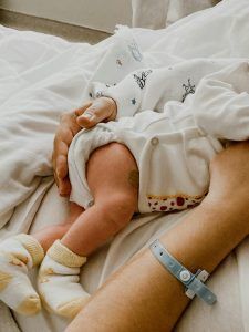 A newborn baby in a diaper and socks, held in someone's arms. The person has an IV band on.