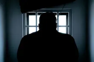 A silhouette of a person standing before a brightly lit window with a grid frame, set against a dark room.