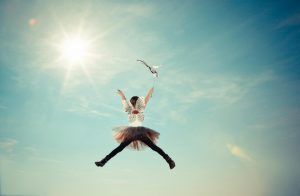 Woman jumping in mid-air, arms reaching for a seagull against a bright blue sky and sun.