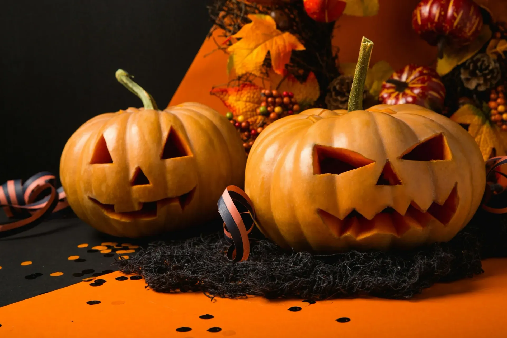 Two carved jack-o'-lanterns sit on a surface with orange and black colors, decorated with fall leaves and confetti.