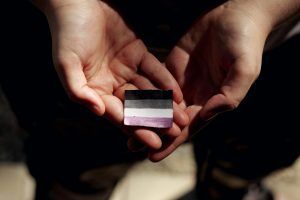 Hands holding a small square with the asexual pride flag: black, gray, white, and purple stripes.