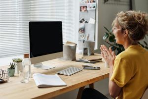 Woman at desk with computer, clapping hands. Yellow shirt, glasses. Wooden desk, blinds, home office.