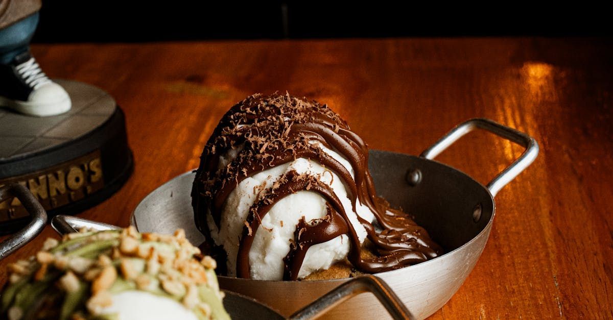 A scoop of vanilla ice cream topped with dark chocolate sauce and shavings, served in a small metal pan on a table.