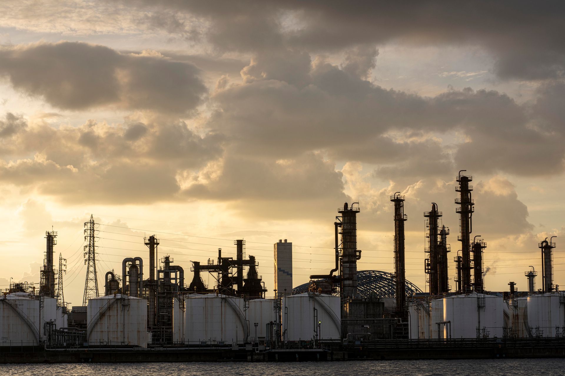 A large oil refinery is silhouetted against a cloudy sky at sunset.