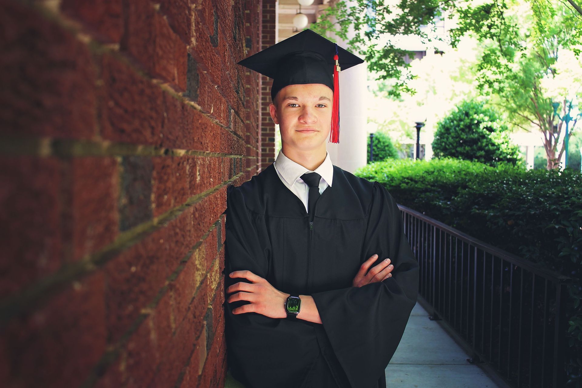 A young man in a graduation cap and gown is leaning against a brick wall.